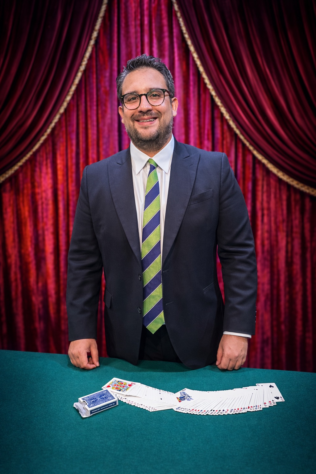 Retrato de Javier Natera junto a una mesa de cartas frente a un telón rojo