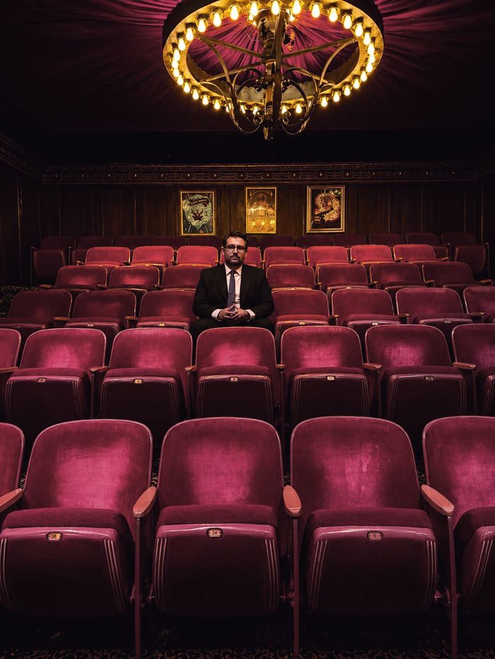 Javier Natera sentado solo en un teatro vacío de butacas rojas, bajo un candil encendido